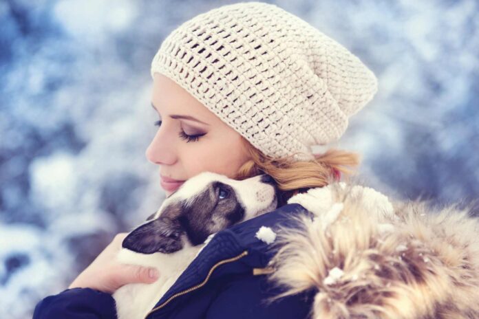 A woman dressed in cold weather gear holding her puppy on her shoulder to keep it warm.